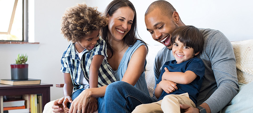 Happy multiethnic family sitting on sofa laughing together. Cheerful parents playing with their sons at home. Black father tickles his little boy while the mother and the brother smile.